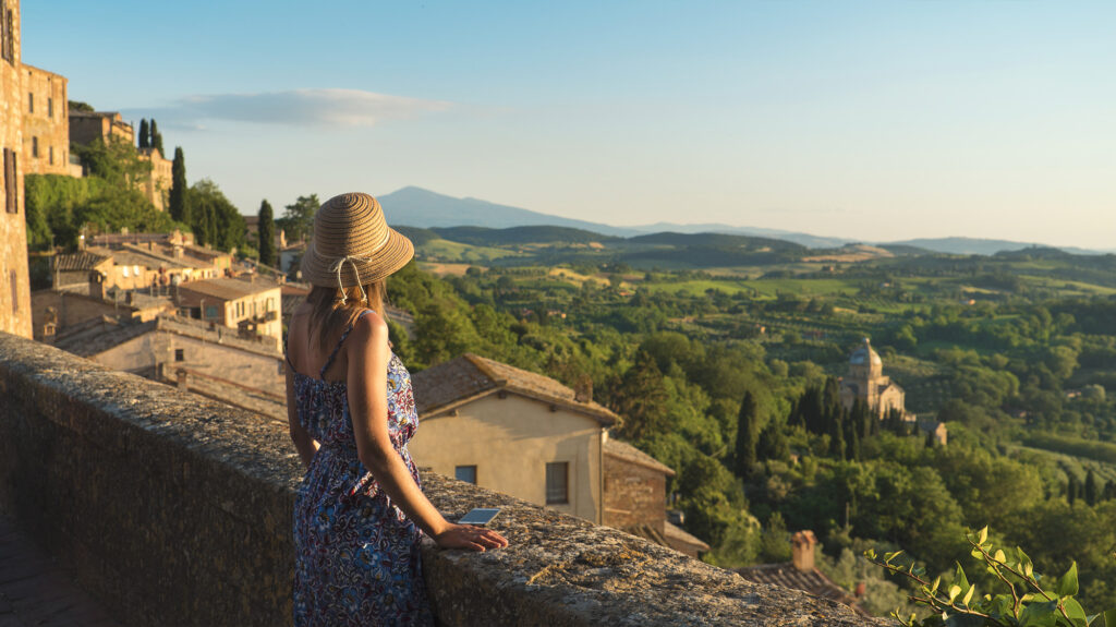 a woman in a sundress and hat looking out over a vast landscape of green trees and fields