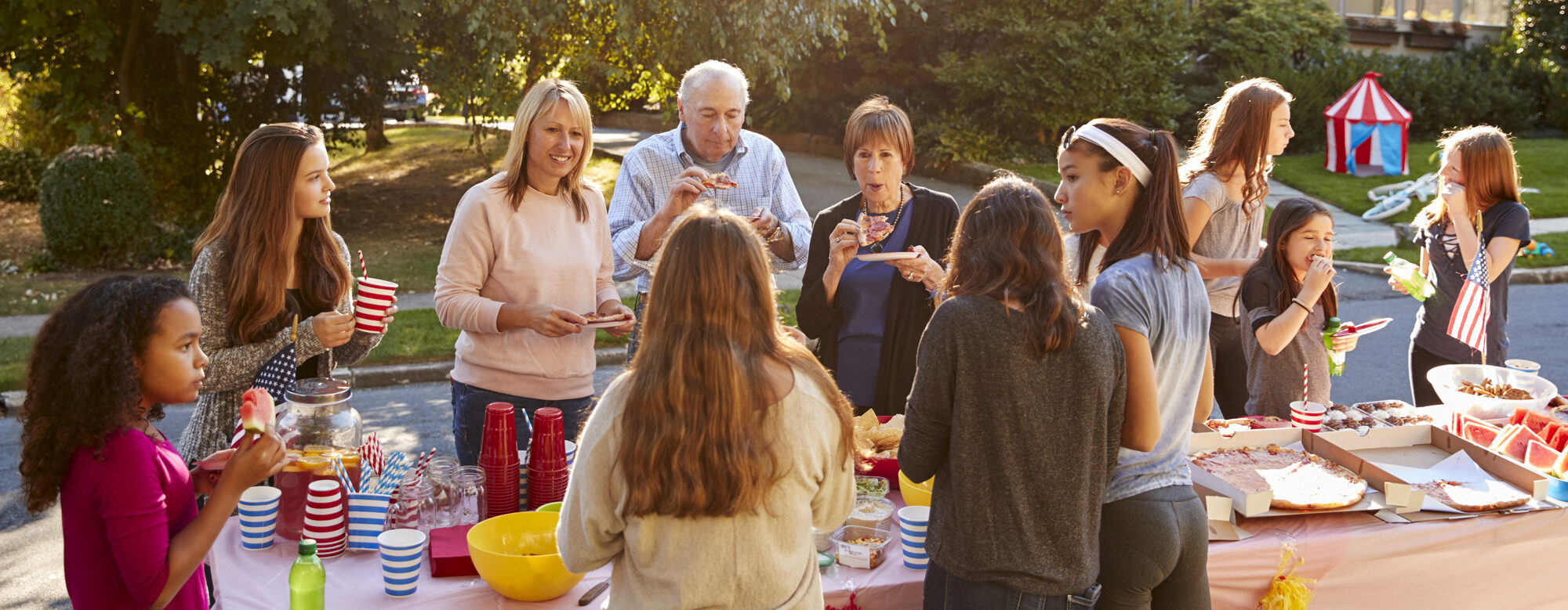an outdoor celebration with a table and food in the street with a diverse group of neighbors of all ages
