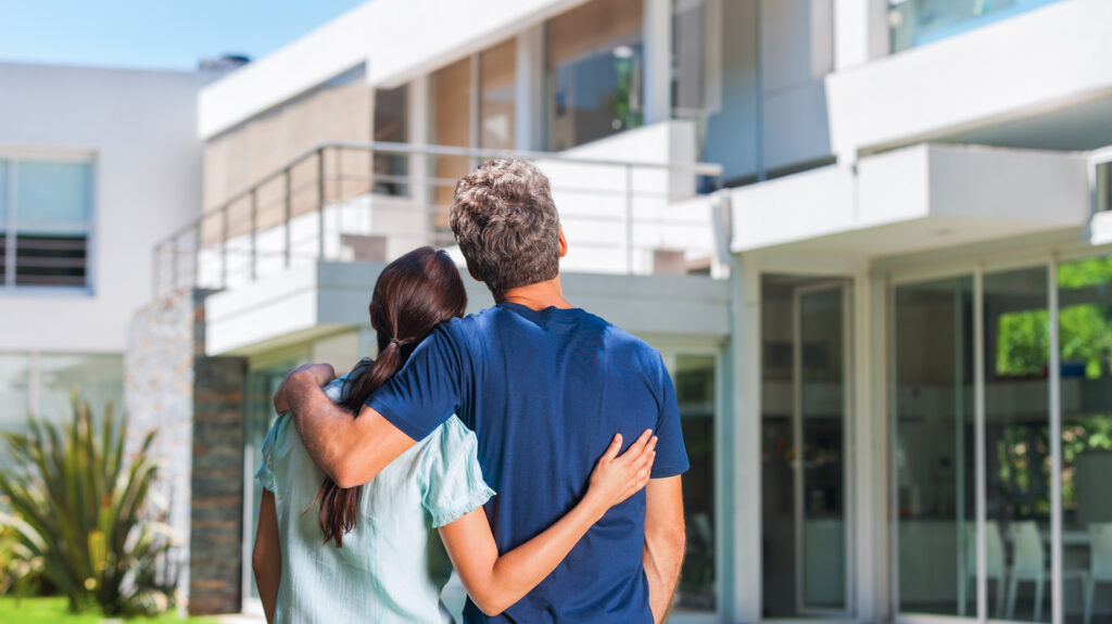 a man and woman in the foreground holding each other around the shoulders looking upon their house which is blurred in the background